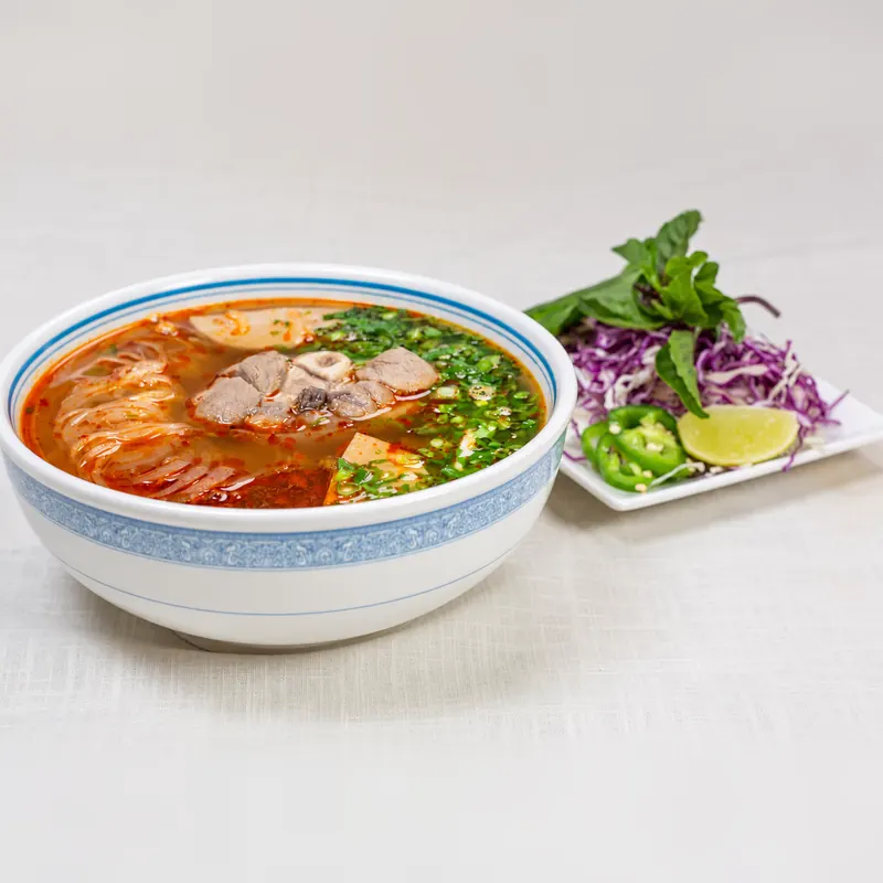 Bún bò Huế in a red chili-lemongrass broth with sliced beef, beside a plate of herbs, cabbage, lime and jalapeño