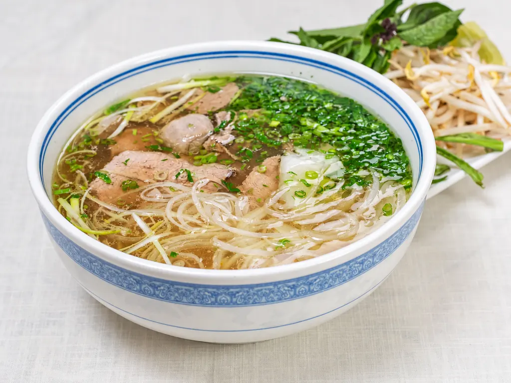A bowl of steak and meatball phở with rice noodles, onion and cilantro, served with bean sprouts, Thai basil, lime and jalapeño
